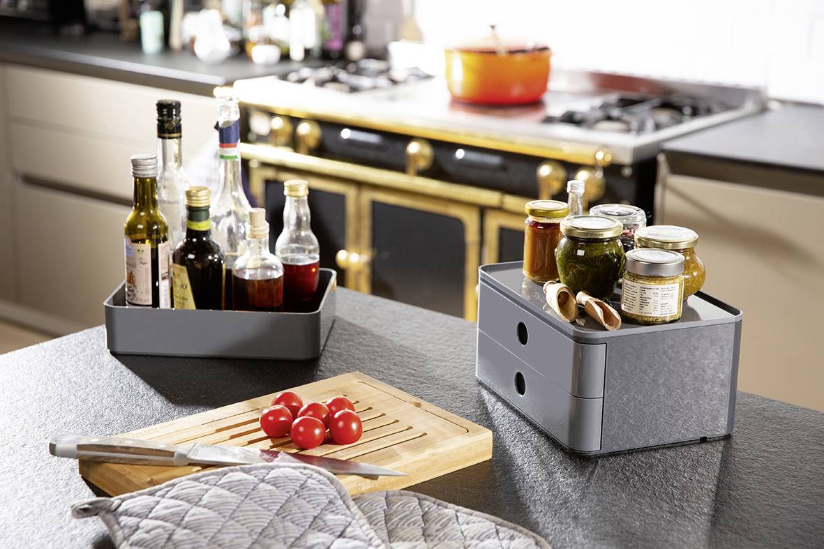 Two grey boxes on a kitchen worktop, filled with spice bottles and jam jars. A chopping board with tomatoes in the foreground.