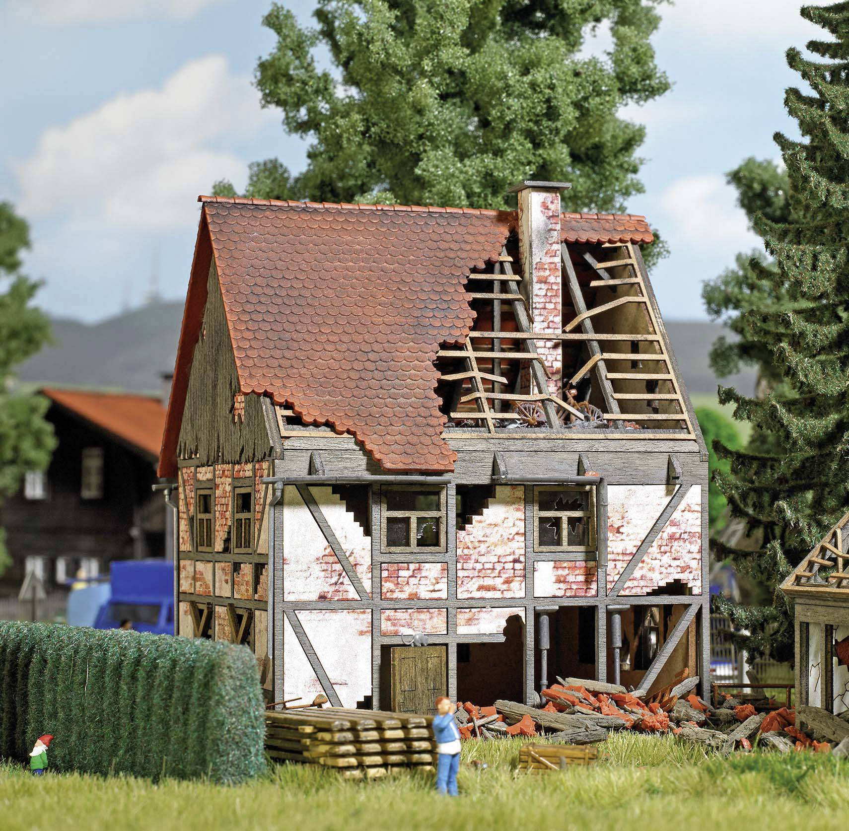 Damaged model house with a broken roof and walls, surrounded by green trees. Two miniature figures stand in front of it.