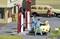 Two miniature figures are filling up a yellow car at a petrol station. Another vehicle with one person is visible in the background.