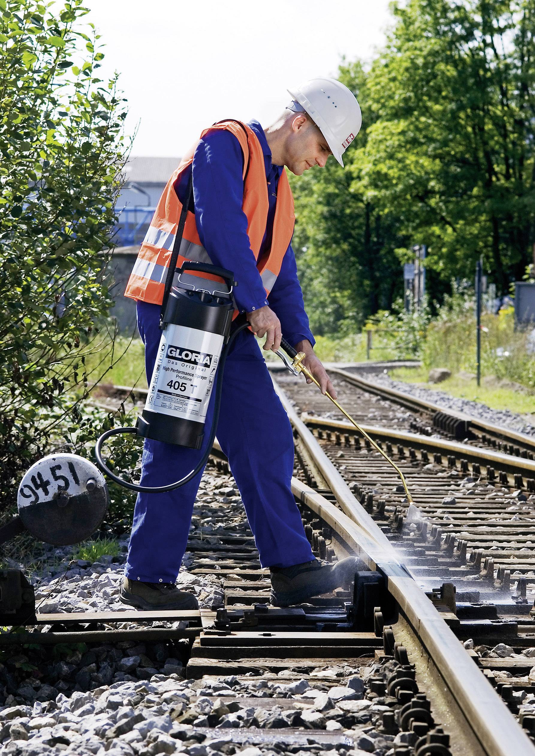 A worker in protective clothing is spraying a substance onto railway tracks for maintenance. Trees and additional tracks can be seen in the background.
