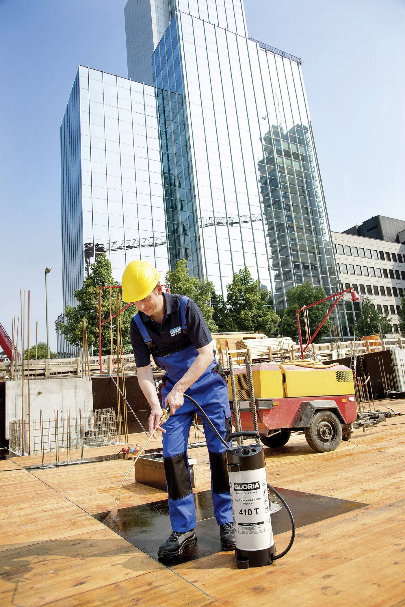 A construction worker wearing a yellow hard hat is spraying liquid onto a wooden board at a building site in front of a modern high-rise building.