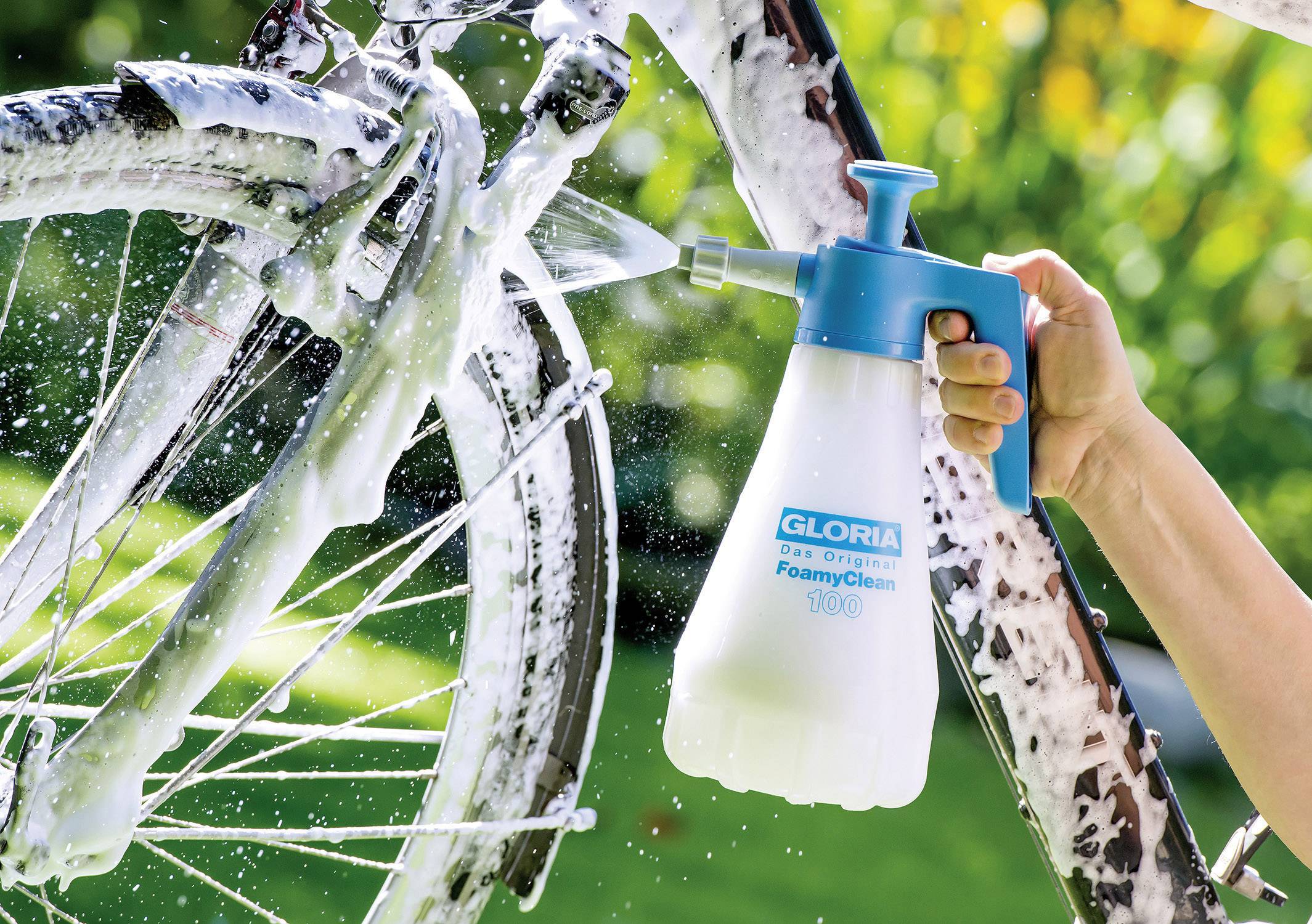 A person is cleaning a bicycle with a blue pressure sprayer, applying soapy water, outdoors on a sunny day.