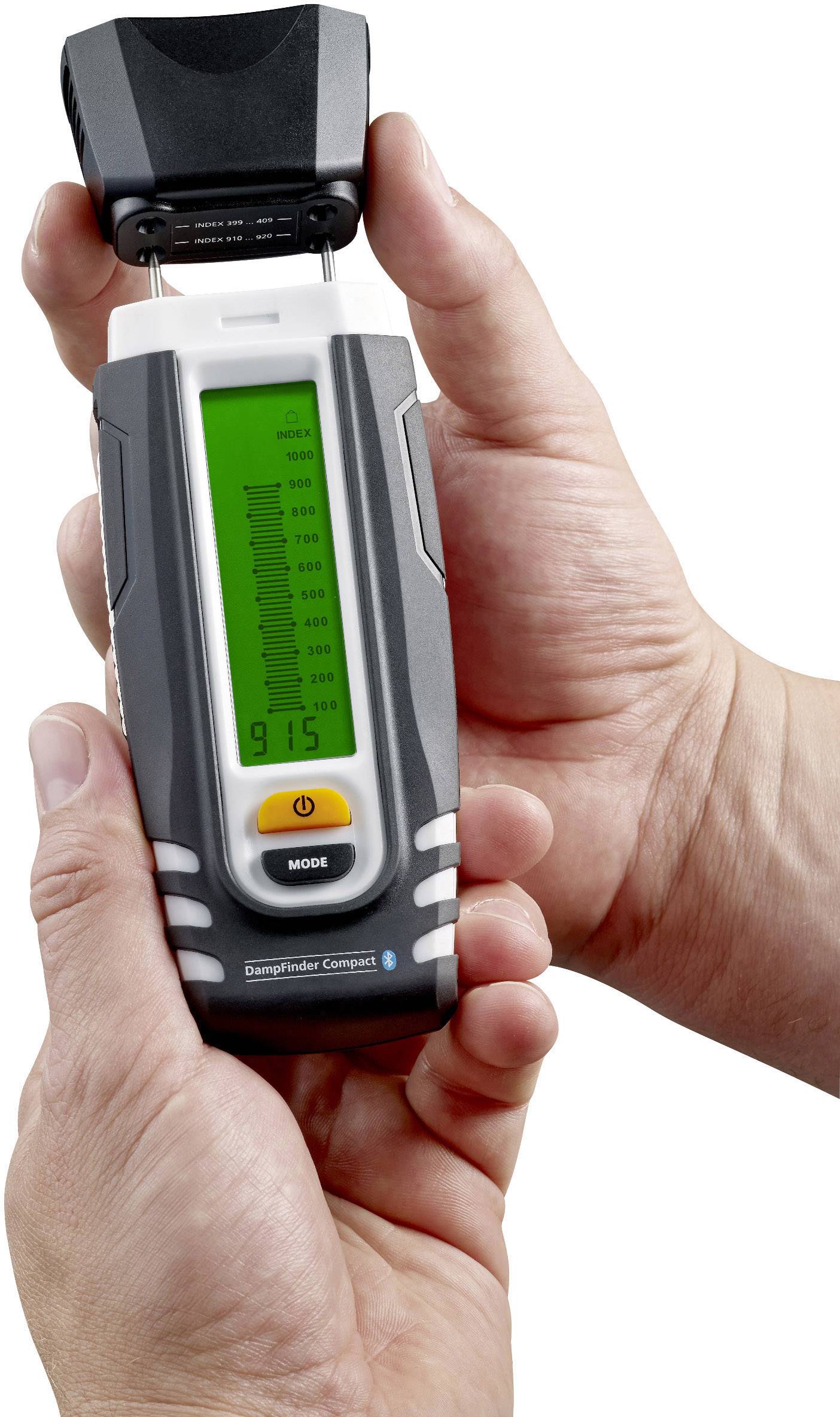 A person is holding a portable measuring device with a green display, showing data. The device has a mode button and a protective cover.