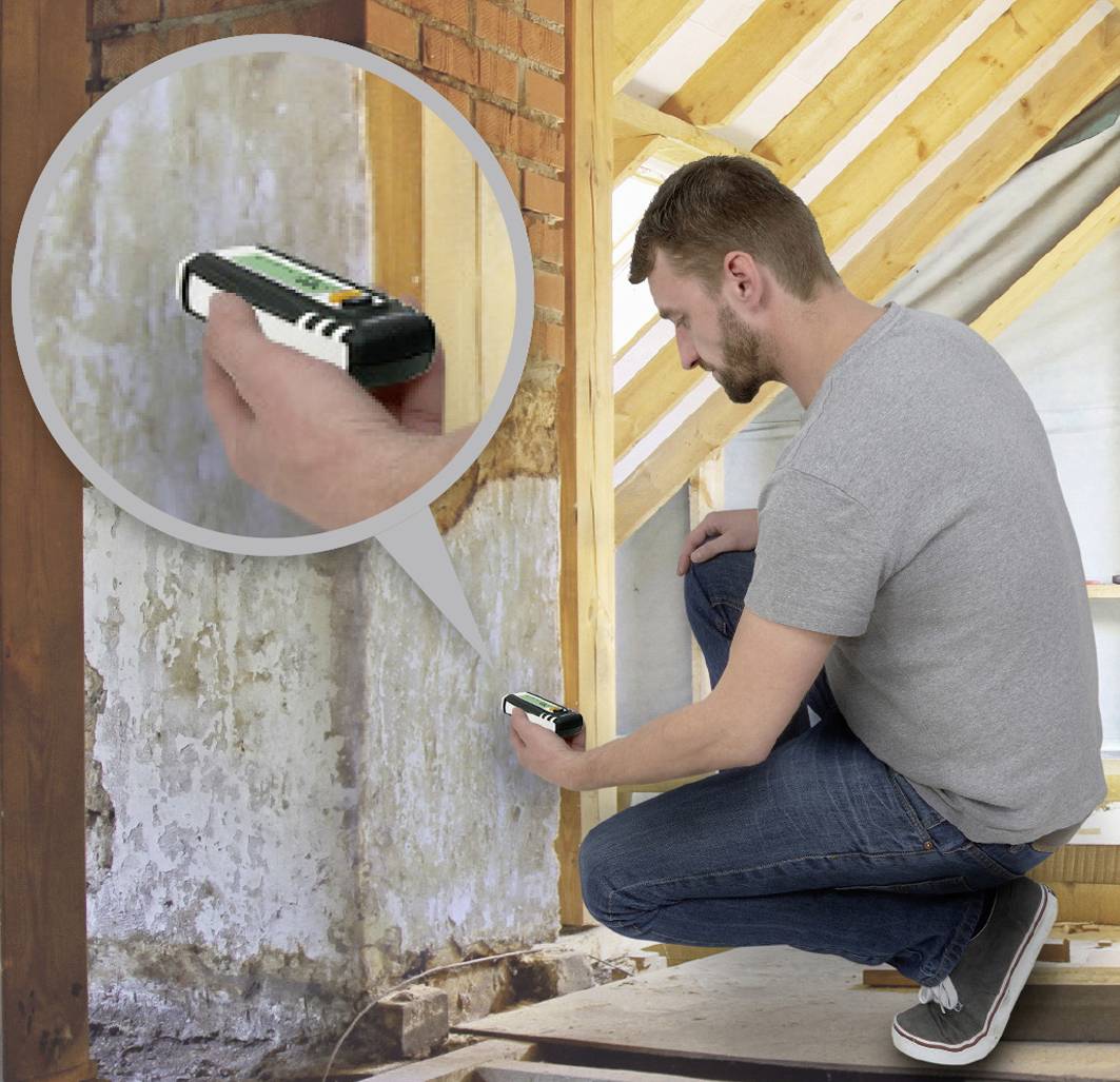 A man is kneeling in the loft and using a moisture metre on a wall to check the moisture content.