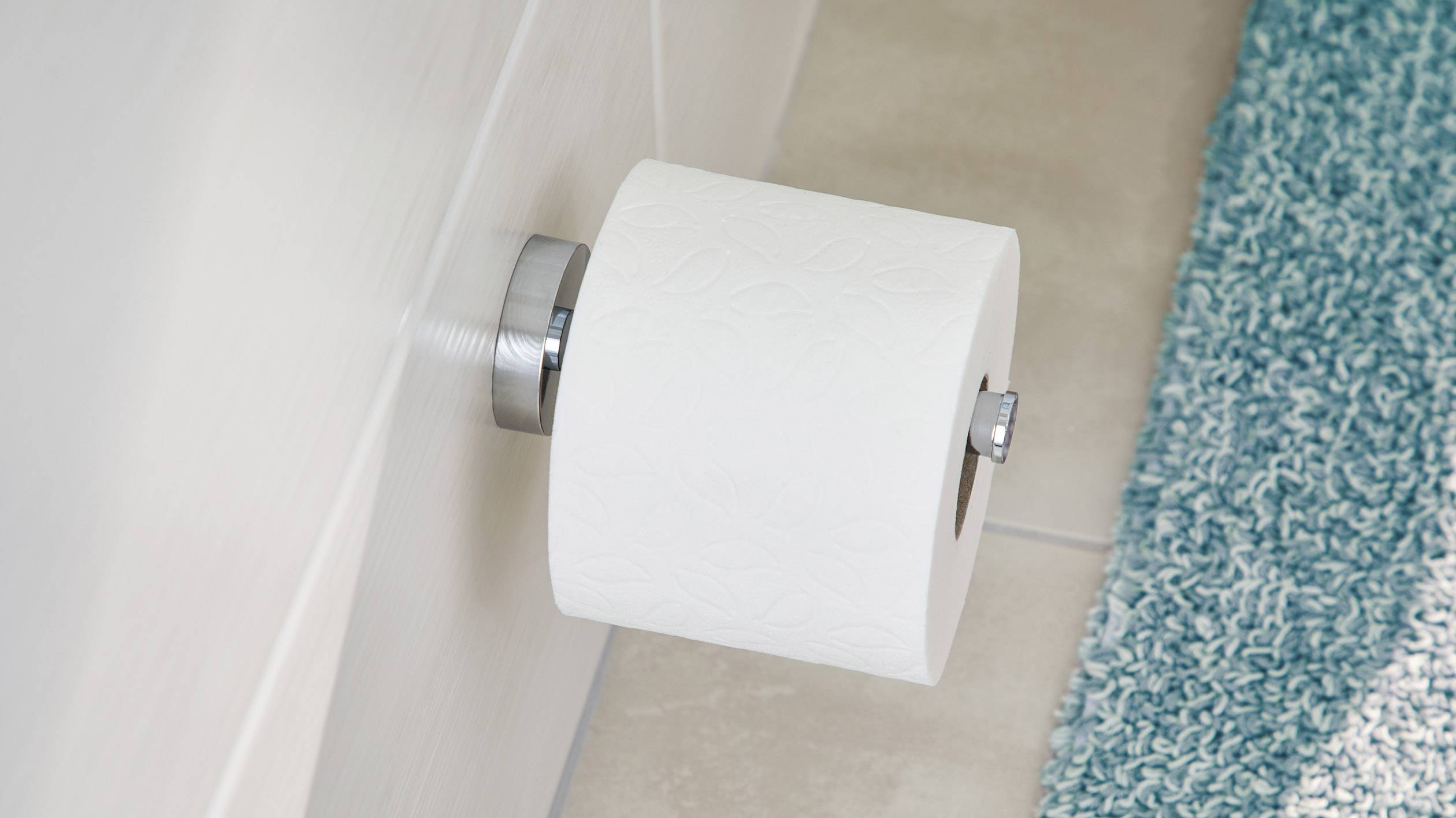 A roll of toilet paper on a wall-mounted holder next to a blue bath mat on a tiled floor.