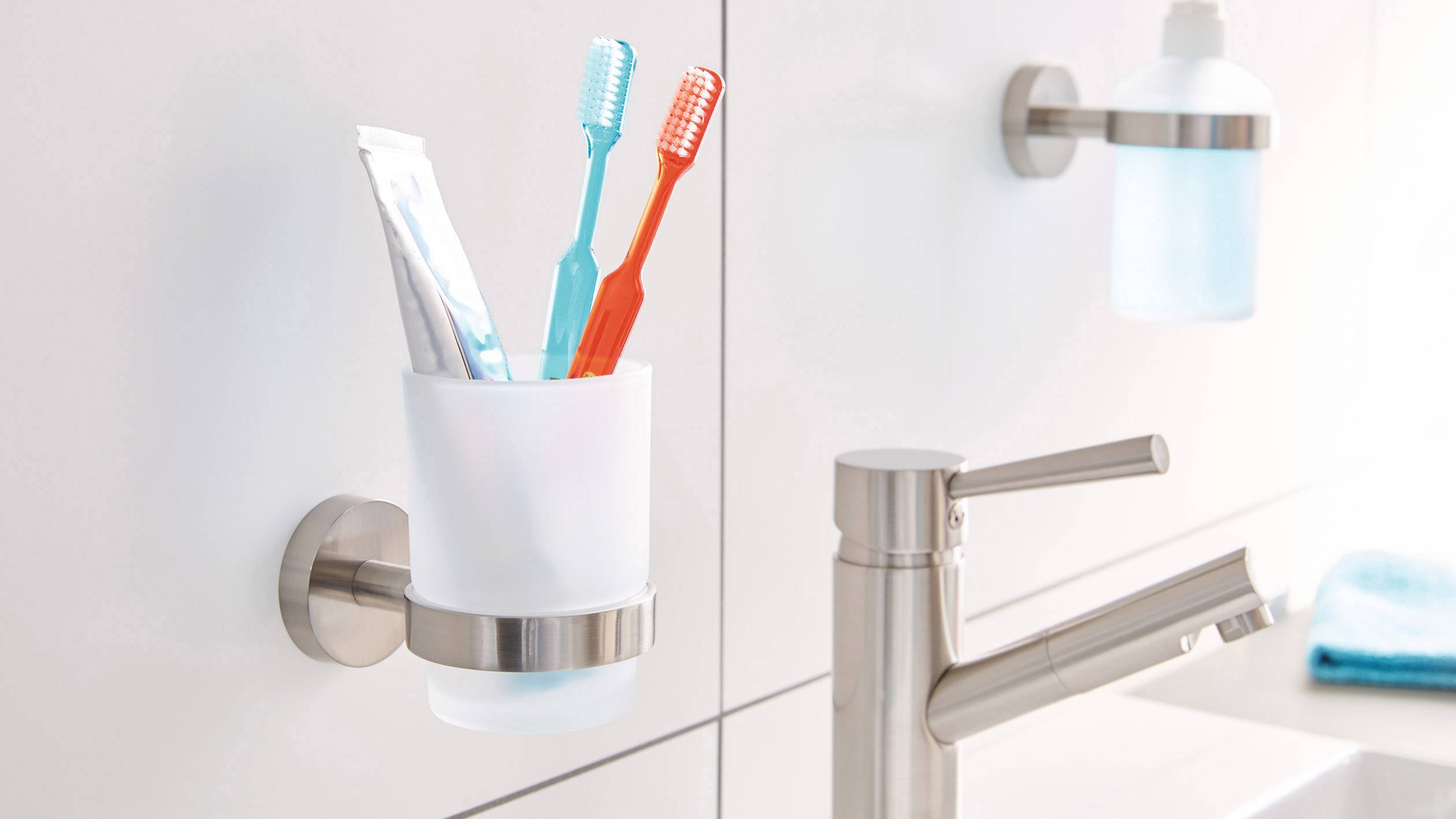 A glass toothbrush holder is mounted on the bathroom wall, holding toothbrushes and toothpaste. Beside it is a tap and soap dispenser.
