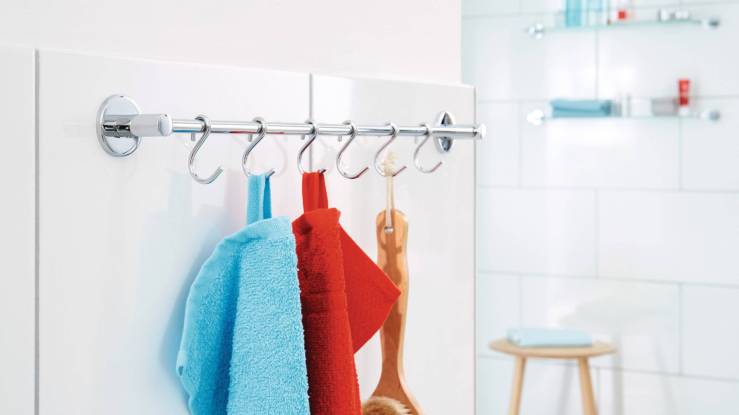 Towels and a brush hang on hooks from a metal rail in a modern bathroom with white-tiled walls and a shelf in the background.