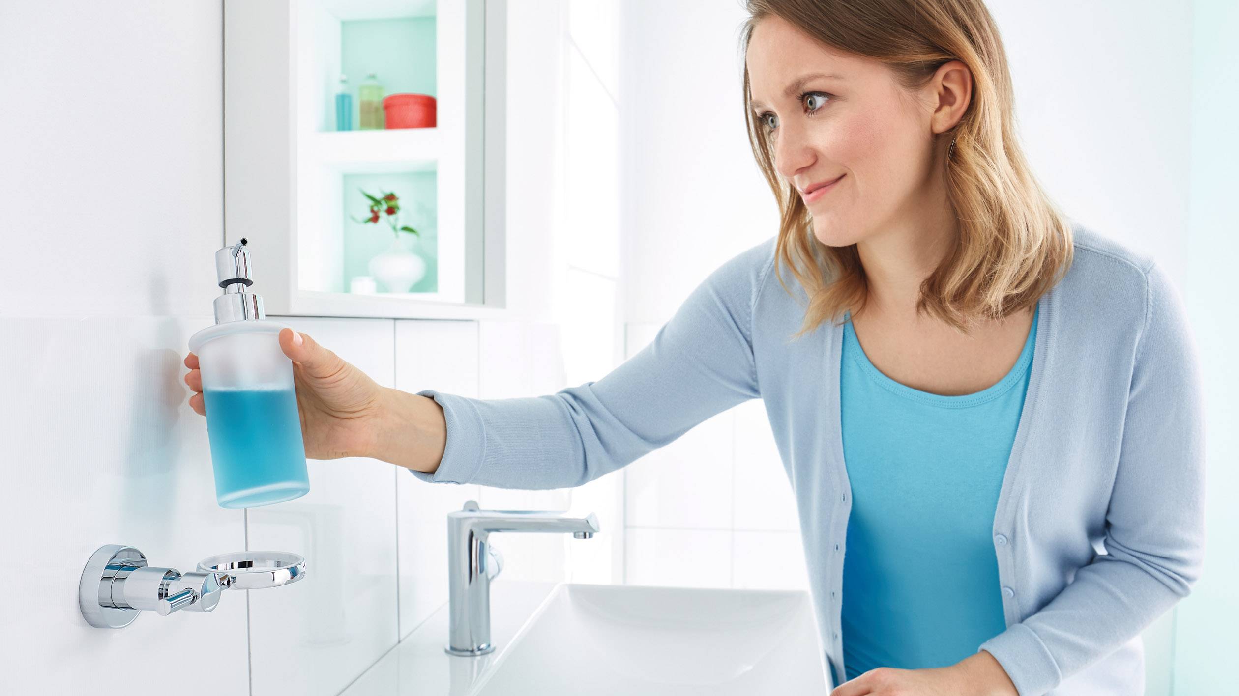 A woman in a bathroom reaches for a soap dispenser next to the washbasin. Shelves with decorative items are visible in the background.