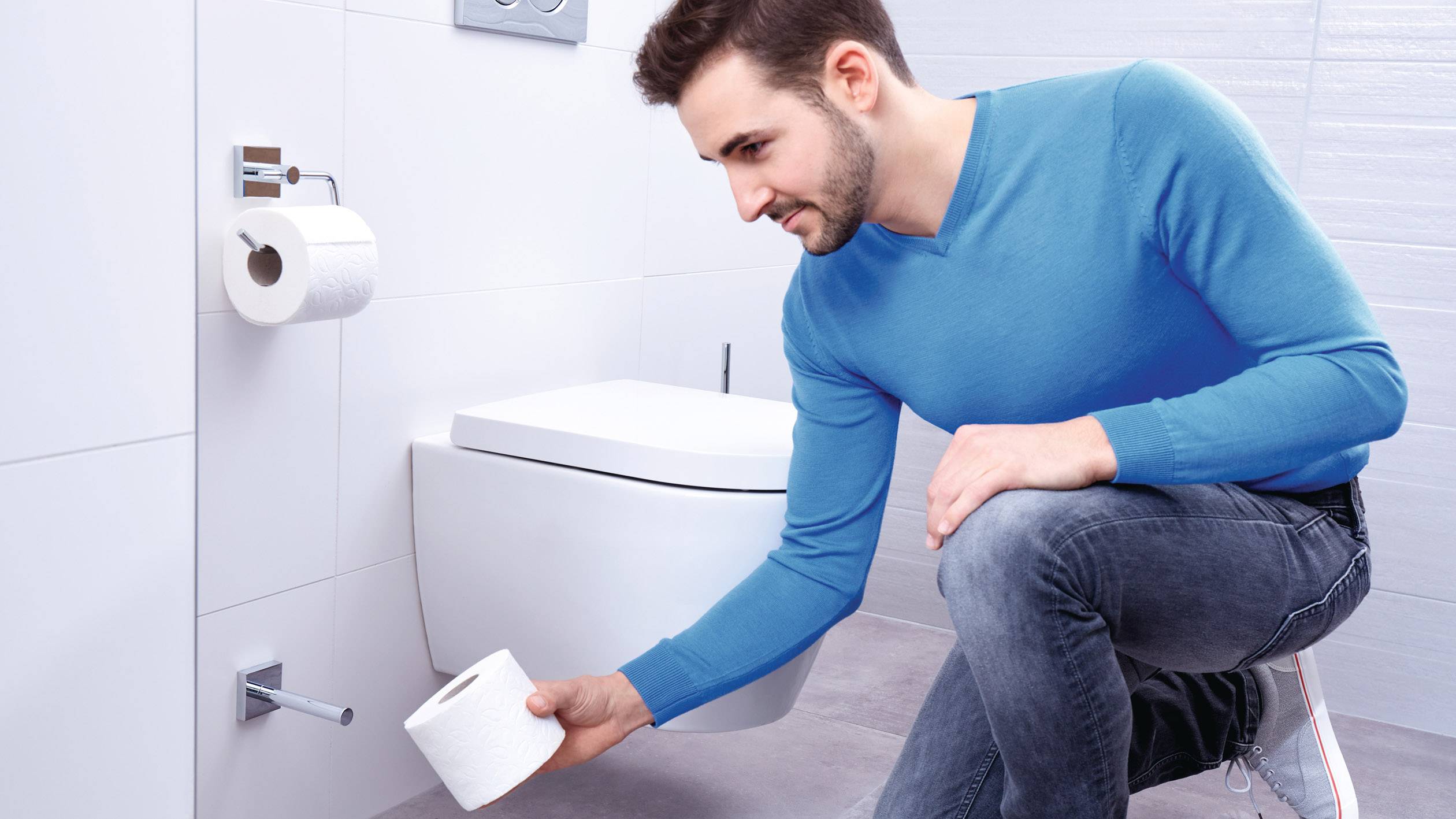 A man in a blue jumper kneels in the bathroom and takes a toilet roll from the holder.