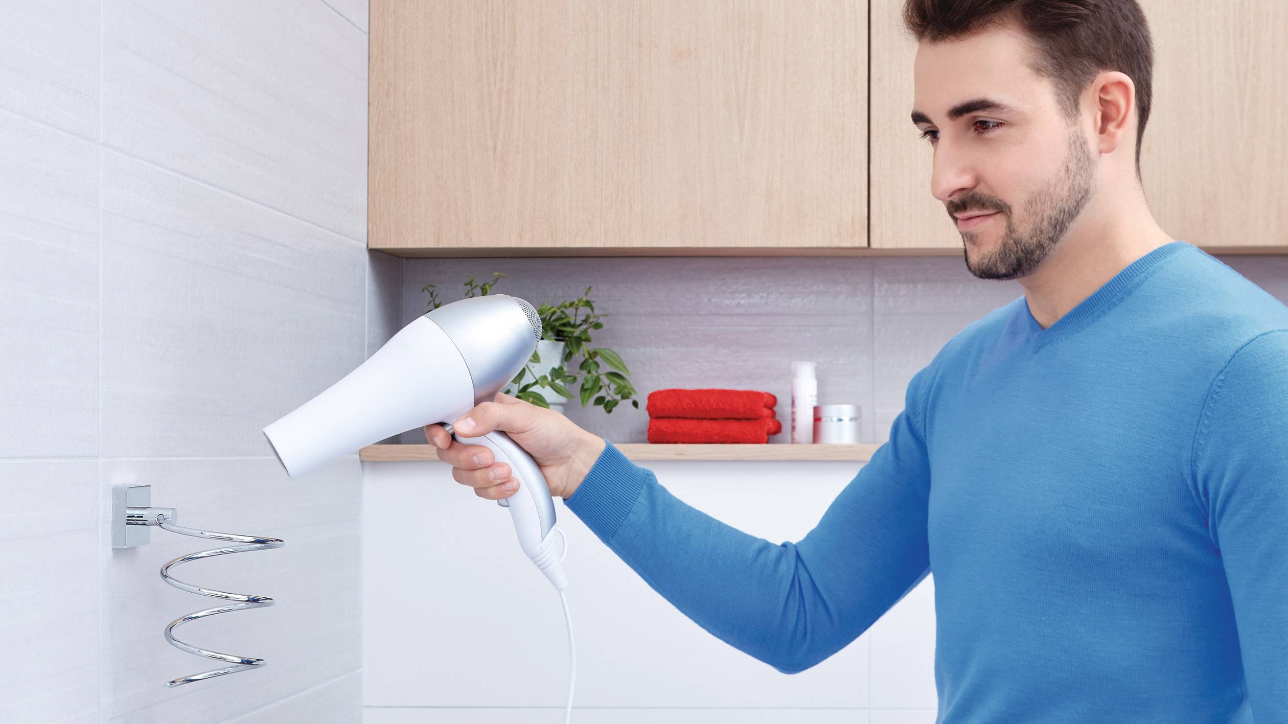 A man in a blue jumper is using a hairdryer on a wall-mounted radiator in a bathroom. Towels and toiletries are visible in the background.