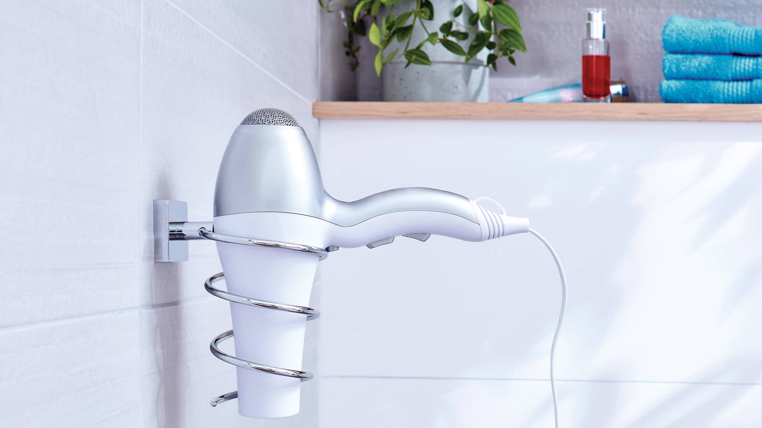 A hairdryer is mounted on the bathroom wall in a holder. Towels and a plant can be seen in the background.