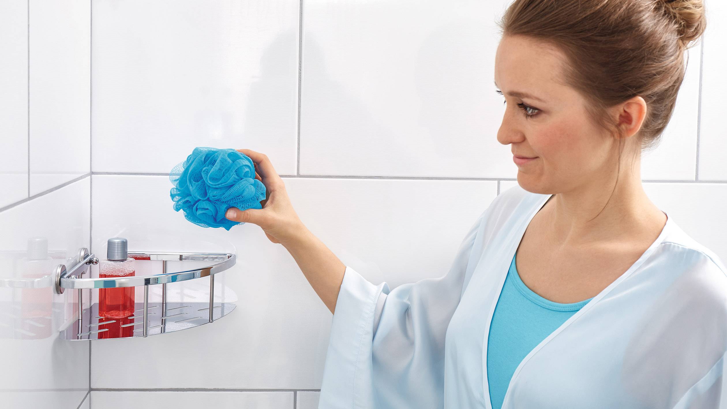 A woman in a blue bathrobe is holding a blue shower sponge in a tiled bathroom.