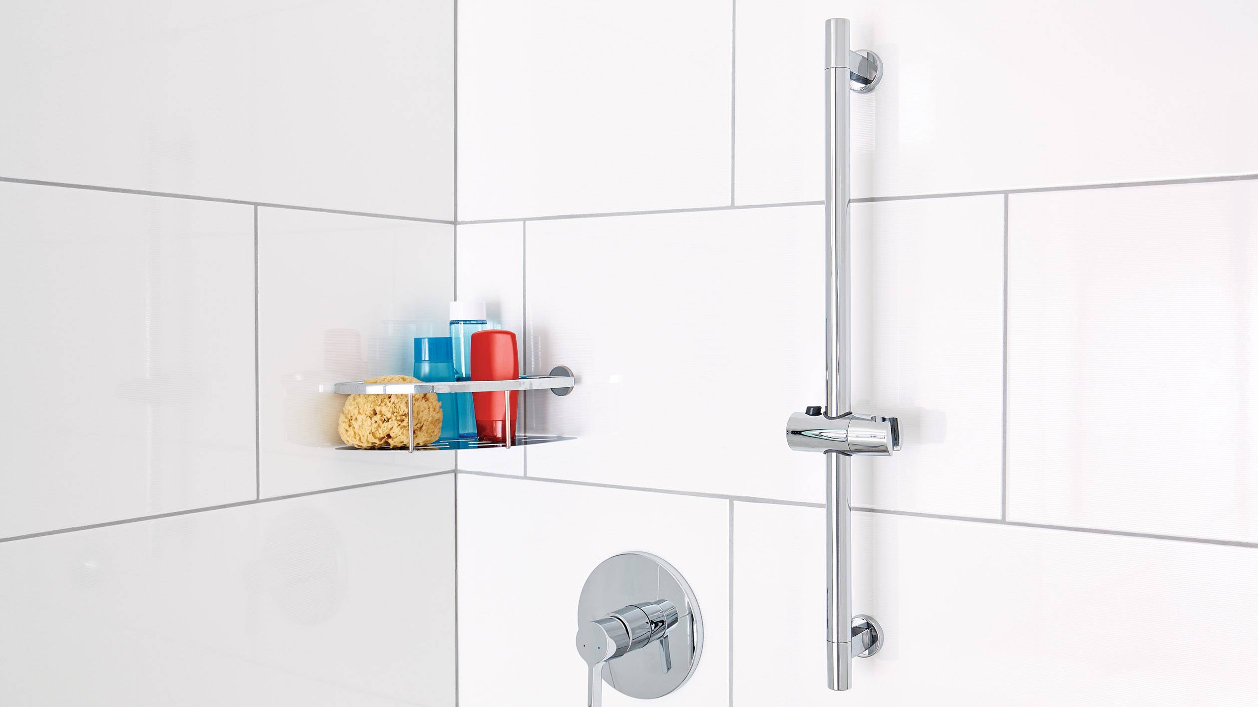 A shower with an adjustable shower rail, controls and a shelf with shower gel, shampoo and a sponge in a white tiled environment.