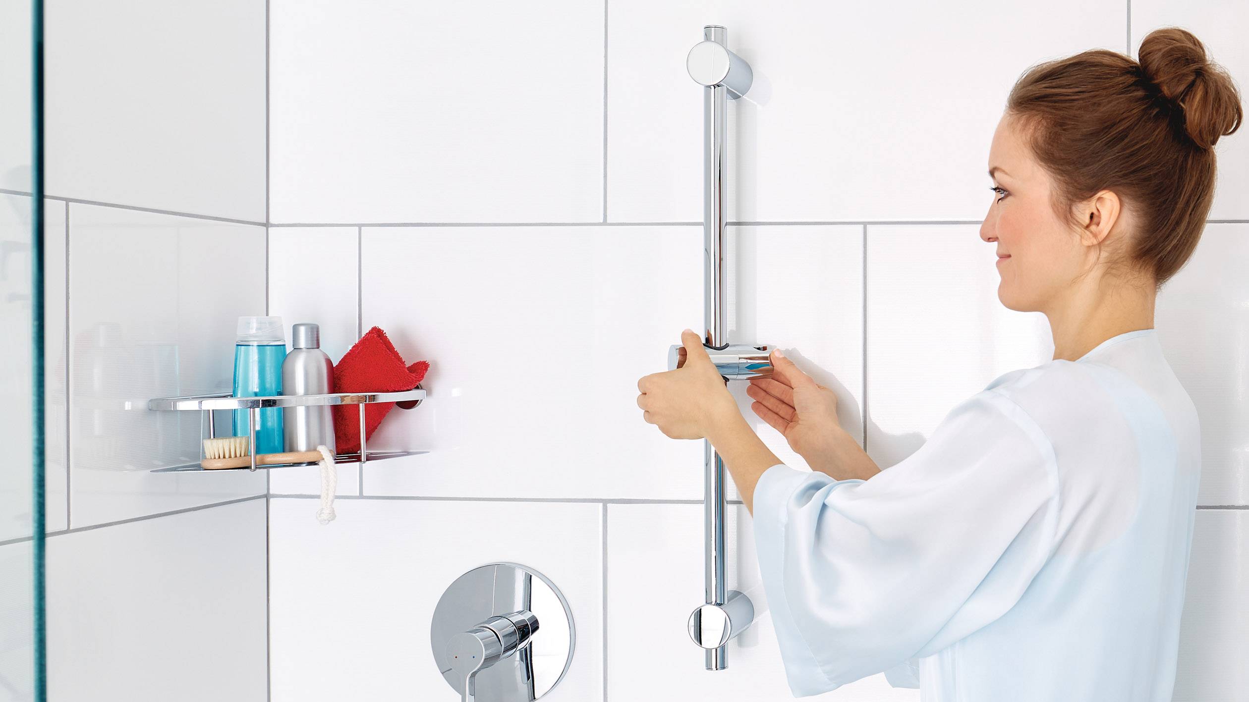 A person in a bathrobe adjusts the shower head mount in a modern bathroom. Shampoo and other toiletries are visible in the background.