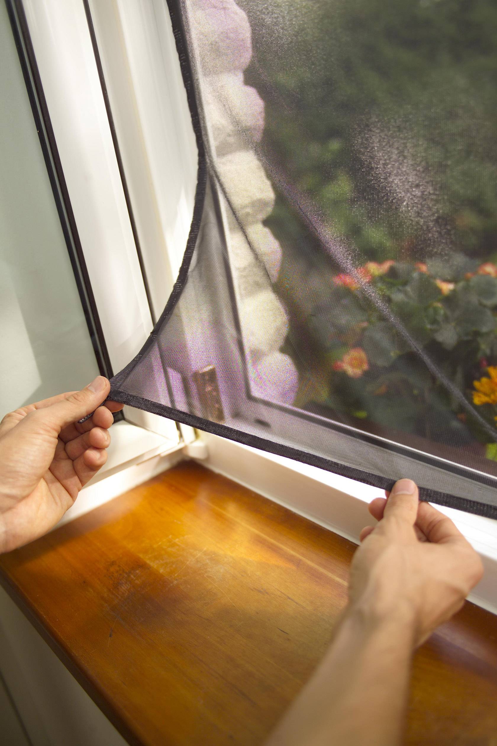 A person is attaching an insect screen to an open window. Flowers can be seen outside.