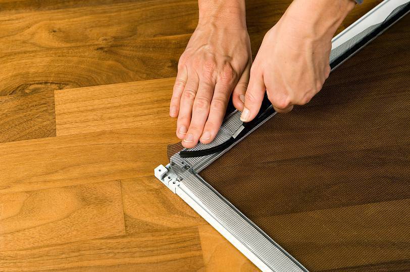 A person is installing an insect screen with a metal frame on a wooden floor by fitting the mesh into the frame.
