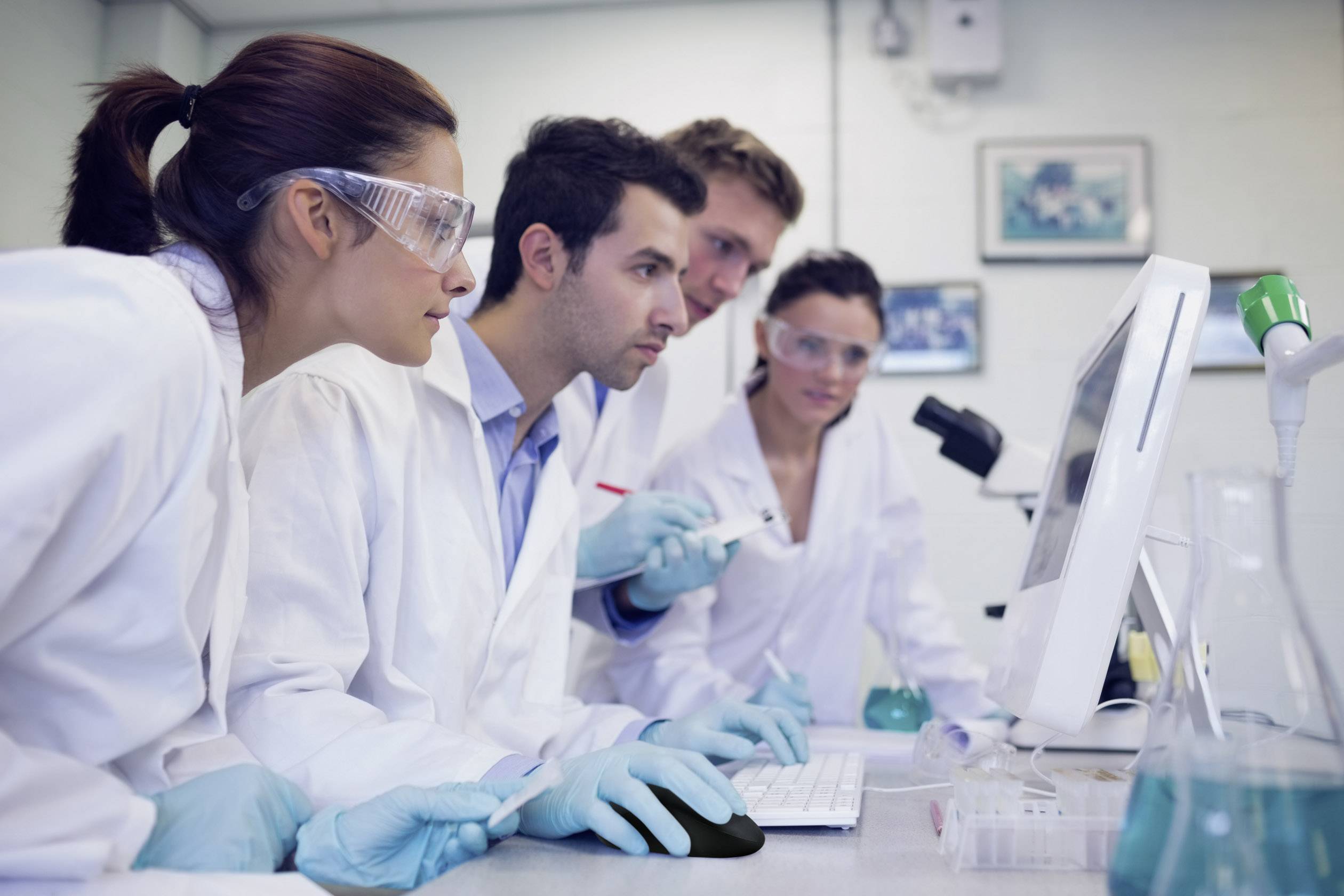 Four people in lab coats and safety goggles are working intently at computers and laboratory equipment in a laboratory.