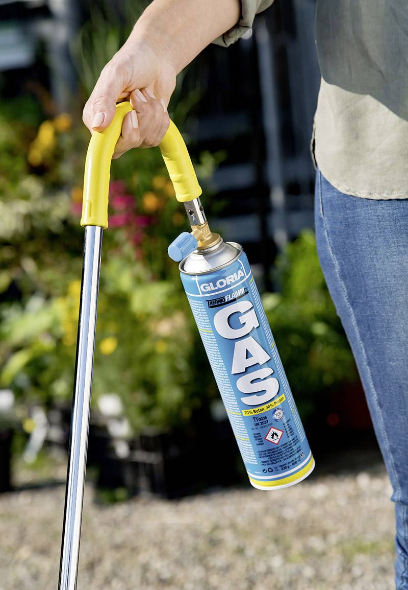 A person is holding a weed burner with a gas canister in the garden. The burner has a yellow handle and is being used to eliminate weeds.