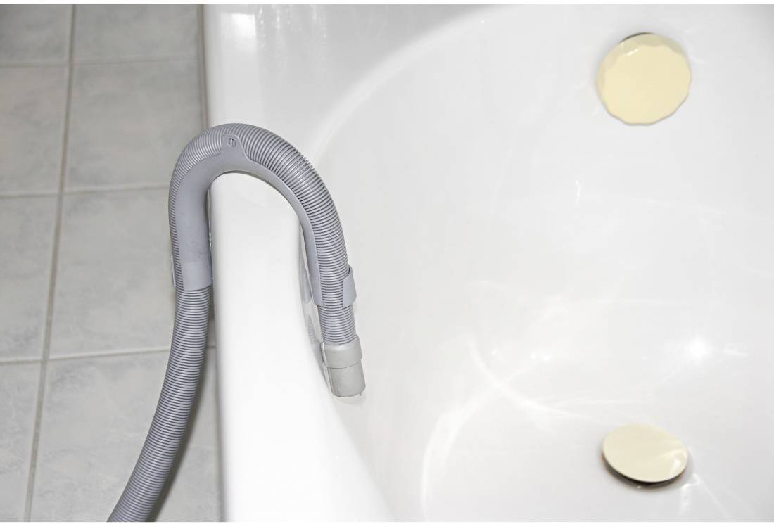 Grey hose hanging over the edge of a white bathtub, next to a gold-coloured overflow and plughole in the bathroom.