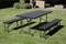 A rectangular table with two benches is situated on a green lawn in front of a modern outdoor area with hedges and furniture.
