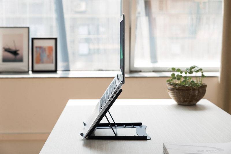 A laptop sits on an adjustable-height stand on a desk. In the background are picture frames and a houseplant.