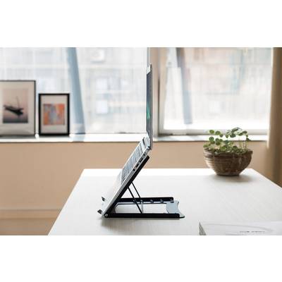 A laptop sits on an adjustable-height stand on a desk. In the background are picture frames and a houseplant.