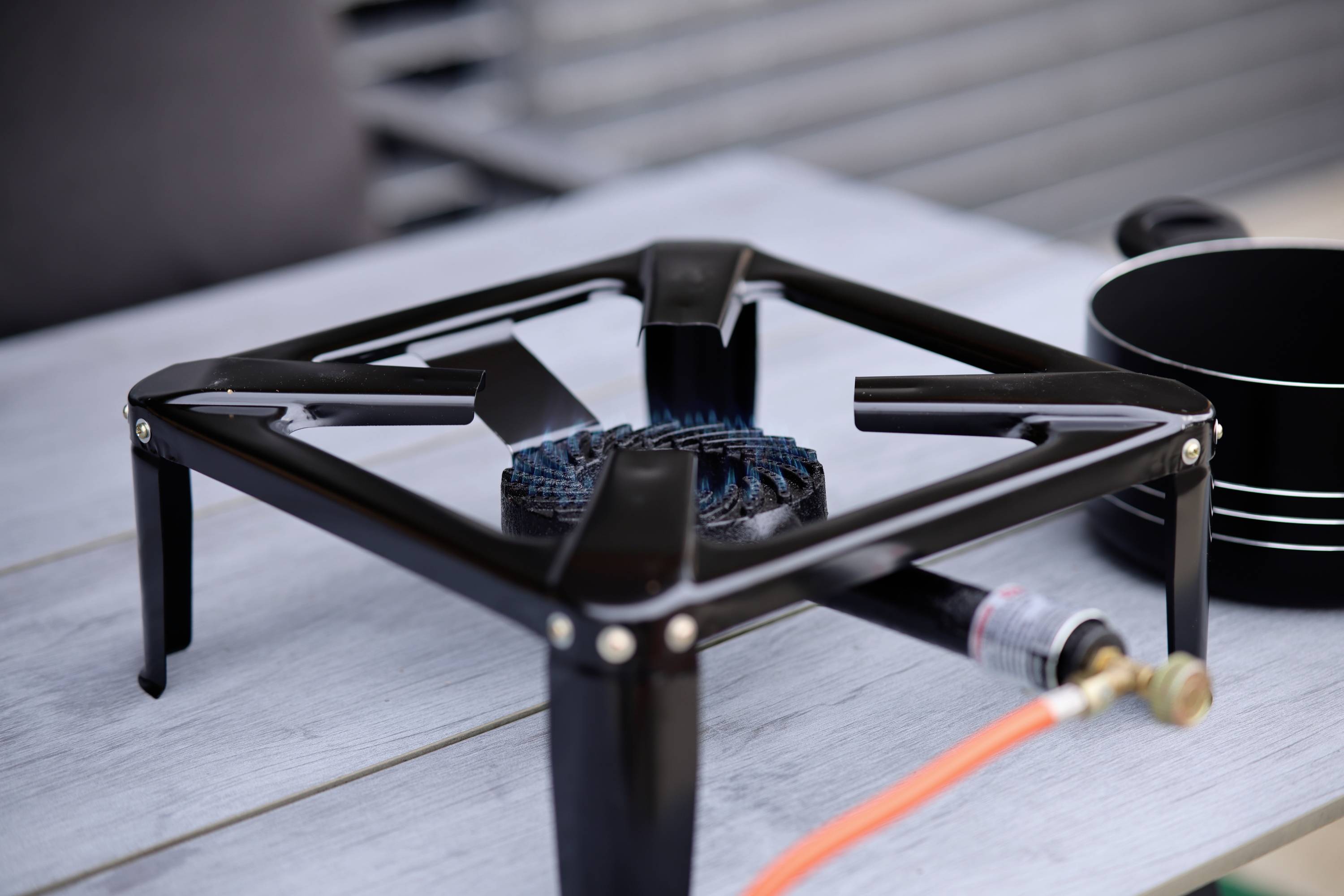 A black gas burner with an orange hose sits on a grey table. Black pots are placed beside it.