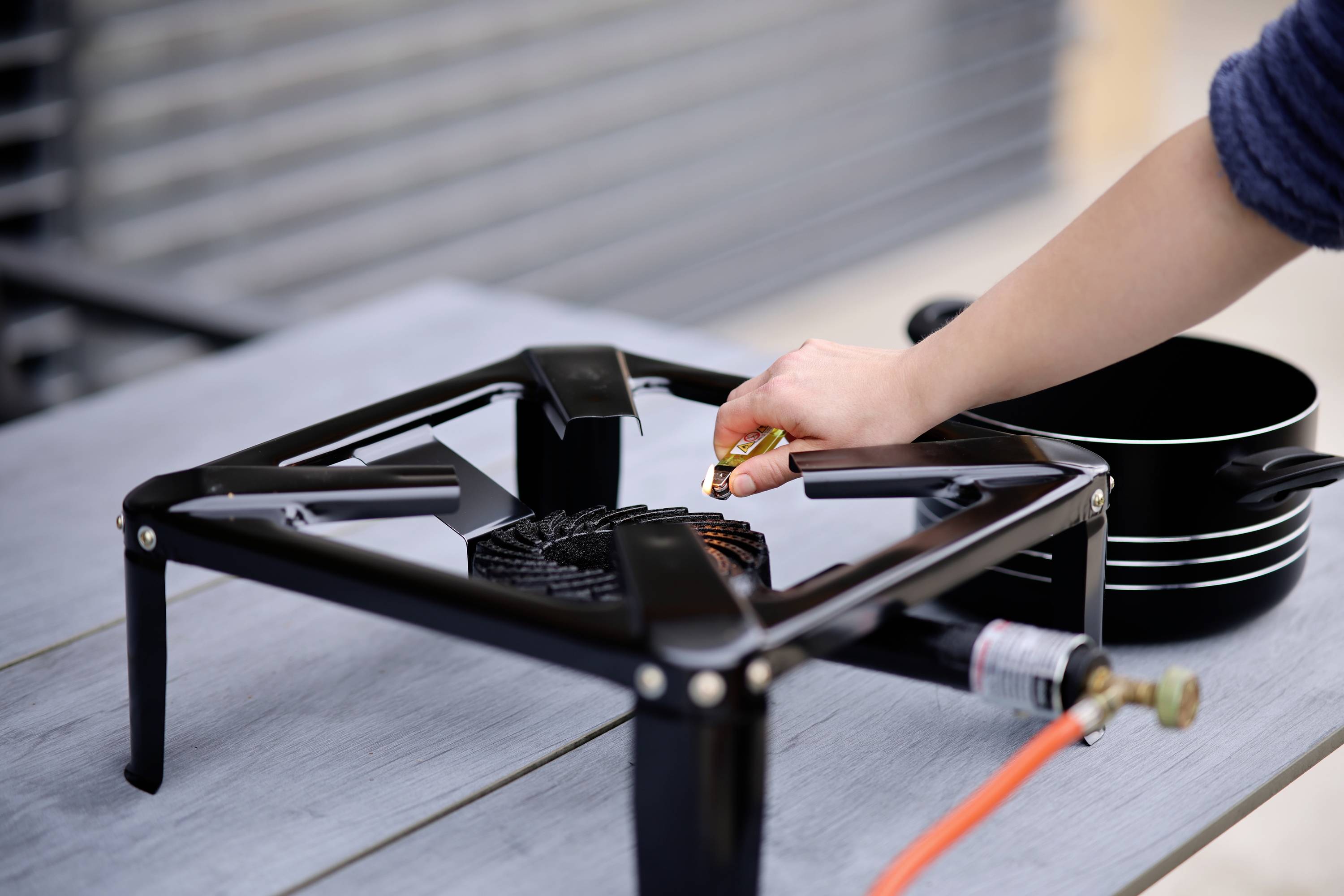 A hand turns a gas hose on a black camping stove on a wooden table outdoors. A saucepan stands ready.