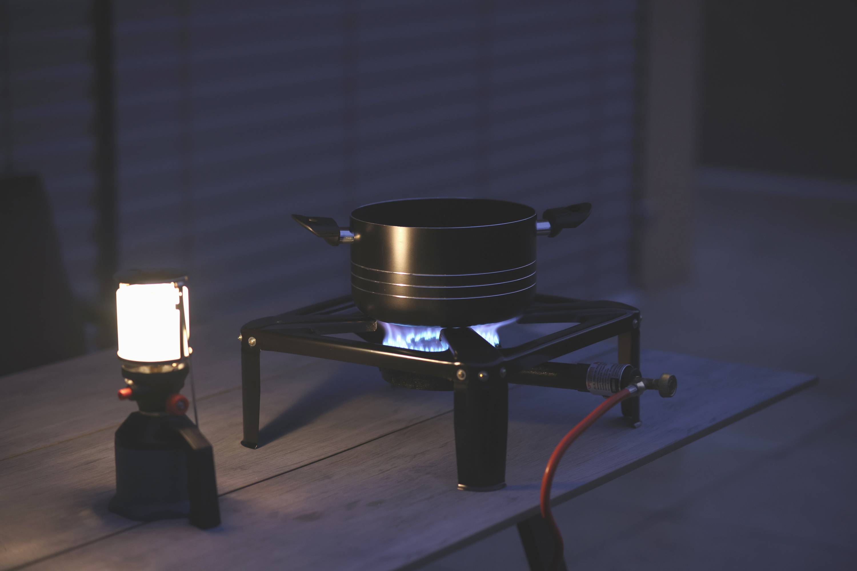 A black pot sits on a burning camping stove on a table. Beside it, a glowing lantern. Twilight in the background.