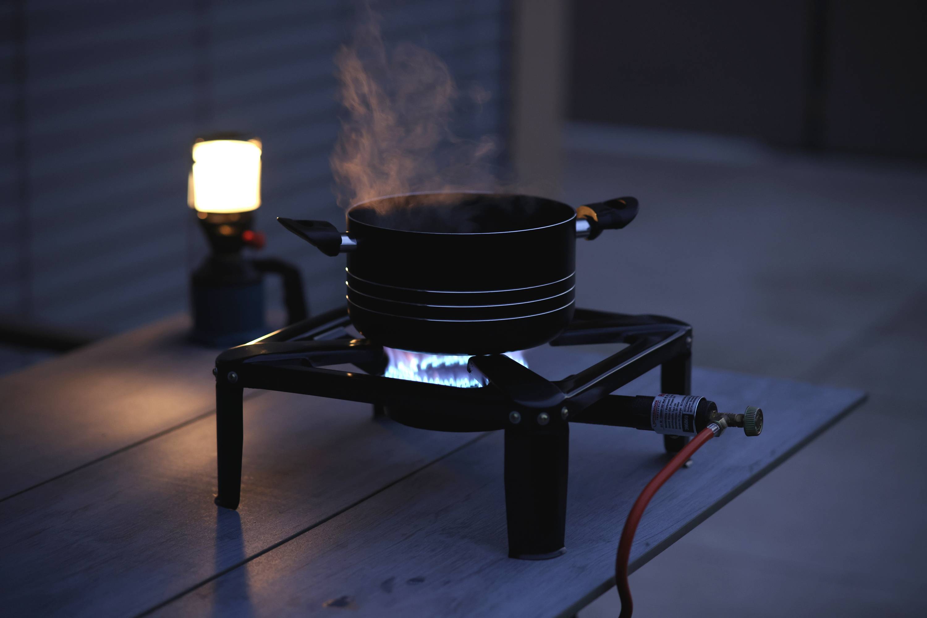 On a table, a pot of boiling water sits on a gas hob. Beside it, a small lamp is illuminated.
