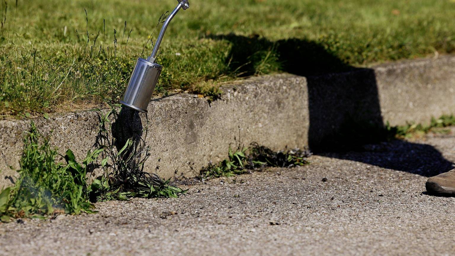 A person is pouring boiling water from a metal watering can onto weeds growing along the edge of a pavement to remove them.