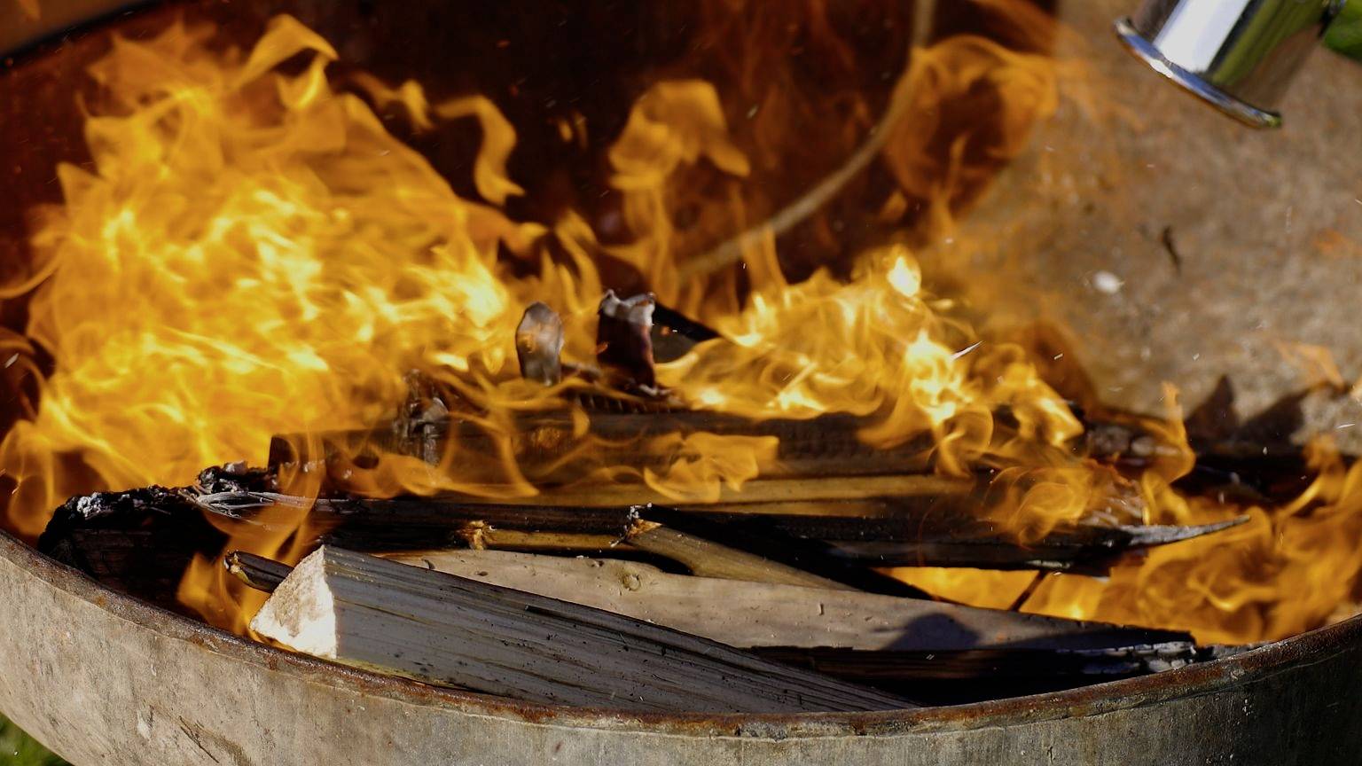 A metal fire basket with burning wood. Flames blaze intensely and wooden pieces glow in luminous orange.