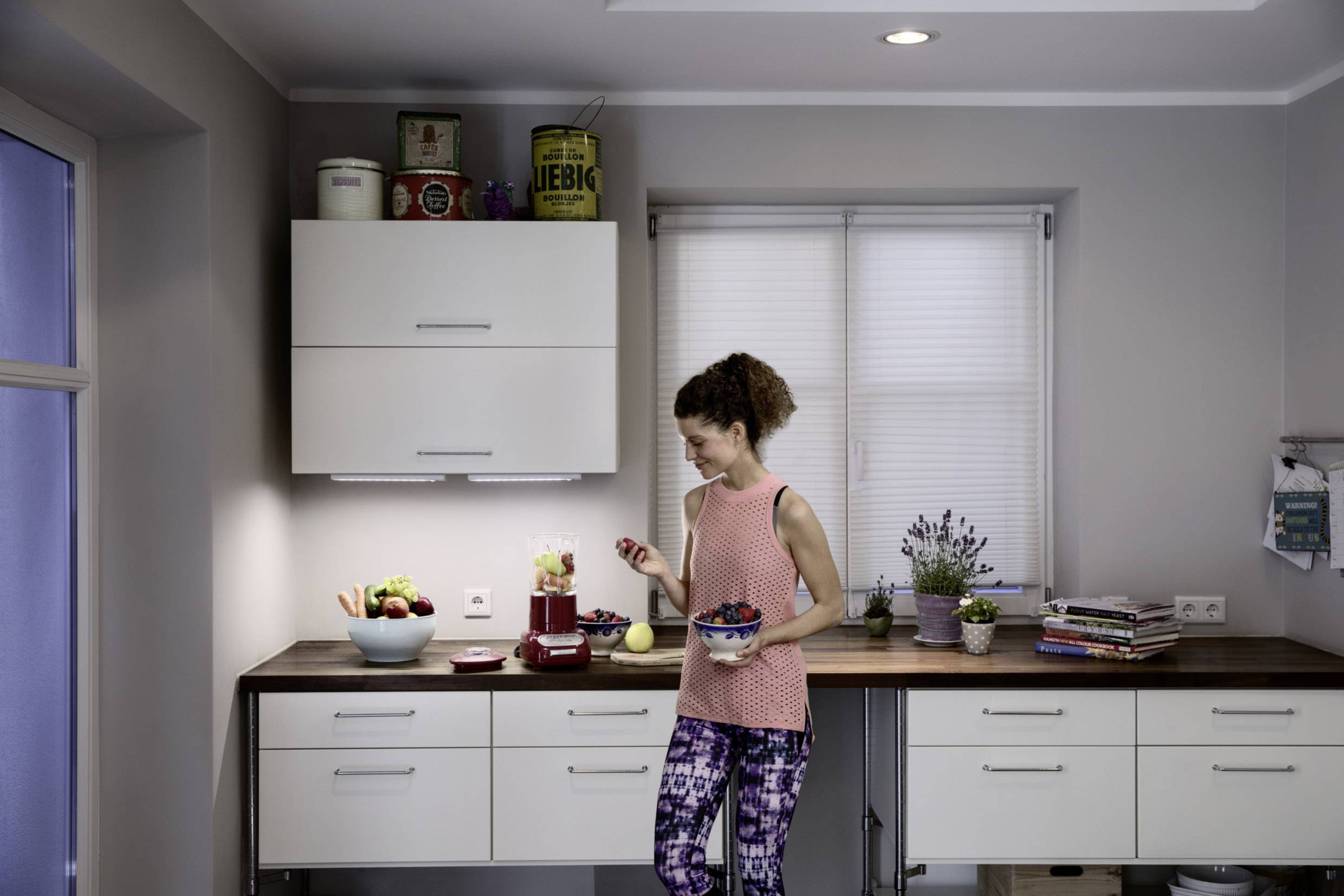 A woman is standing in a modern kitchen, holding a bowl of fruit. Next to her, a blender sits on the worktop with fresh fruit.
