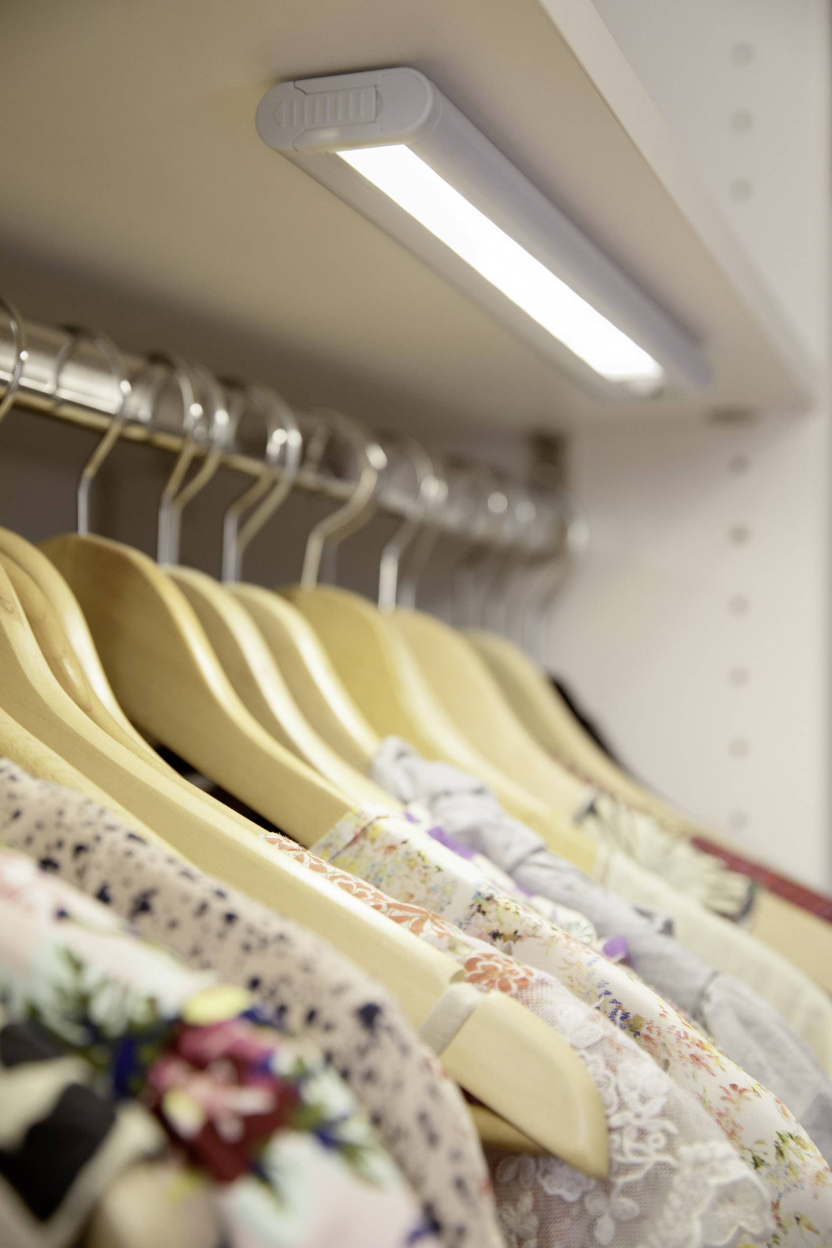 Several wooden coat hangers with colourful clothing hang closely together in an illuminated wardrobe.