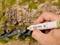 A hand holds a marker and is painting a detailed model of a rocky hill with moss and grass.