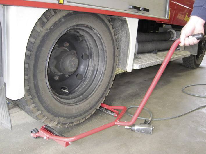 'A red lifting tool is raising a large lorry wheel. One person is holding the handle. Part of the lorry is visible in the background.'