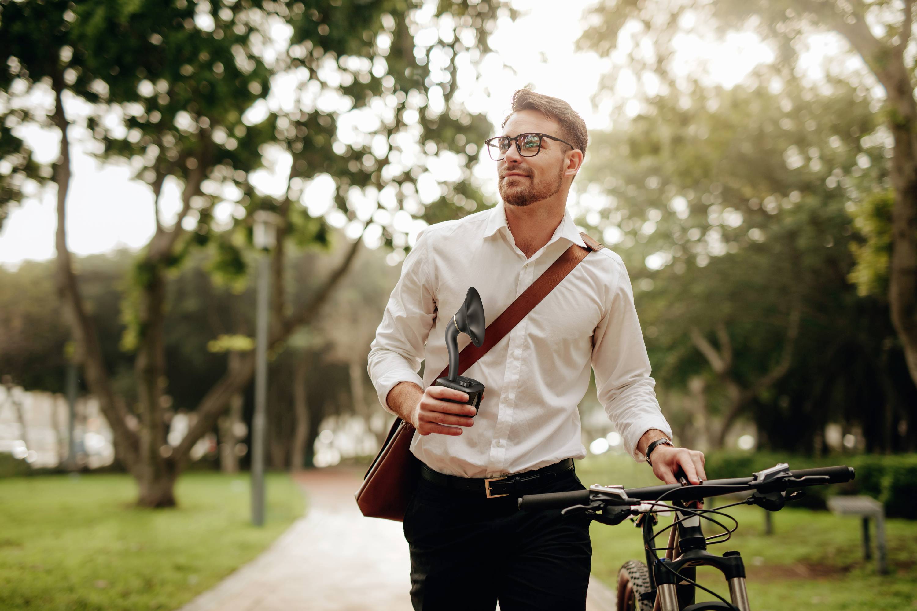 A man in a white shirt is walking along a park path, holding a bicycle and a coffee cup, surrounded by trees and lawns.