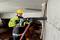 A construction worker in protective gear is drilling a hole in a concrete wall using a drill machine. Construction structures are visible in the background.
