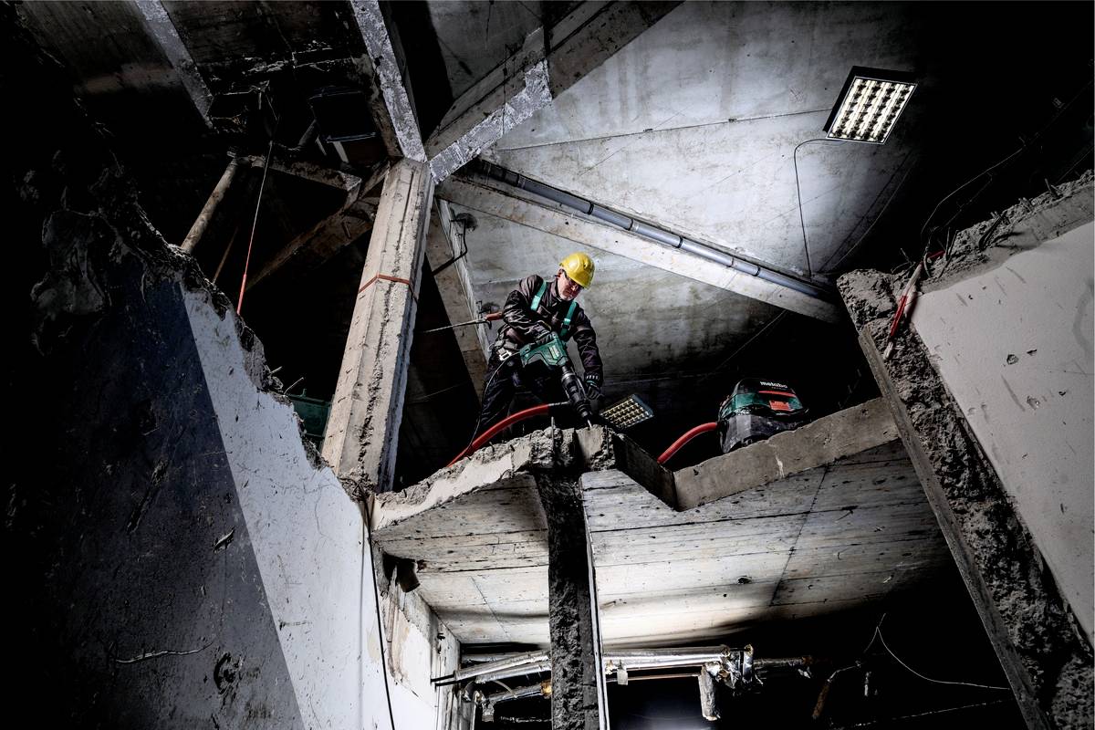 A worker wearing a hard hat stands on a building site with concrete debris, using a tool to drill or cut through concrete.