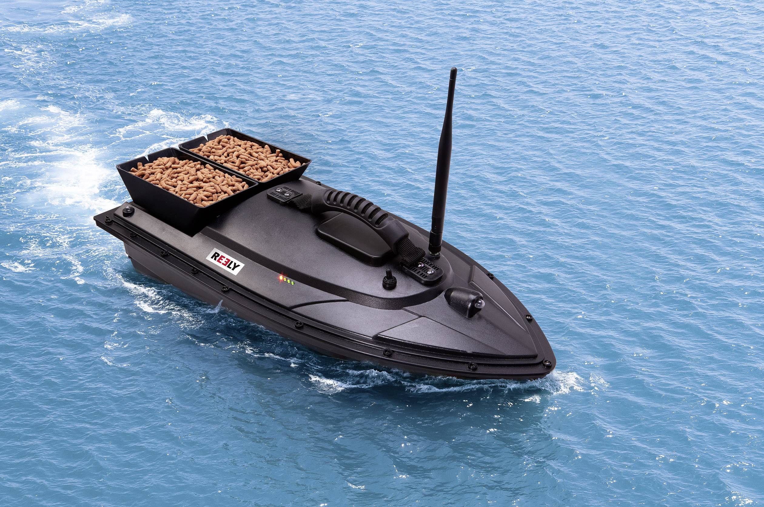 A remote-controlled black boat on water, with two open compartments filled with brown pellets. The boat has an aerial.