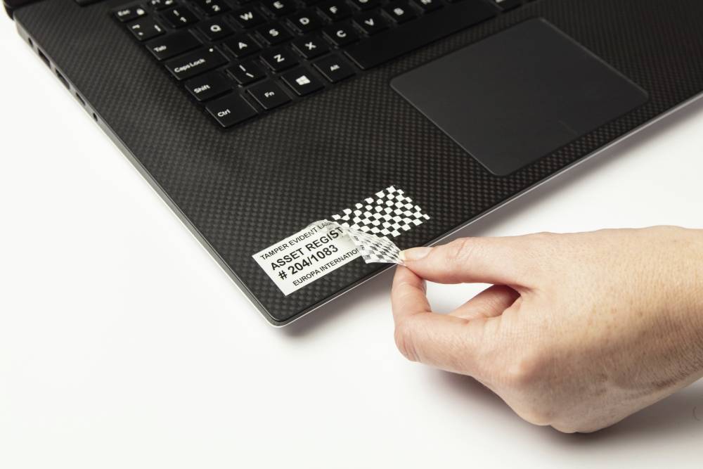 A hand removes a security sticker from a laptop. The sticker displays a serial number and a diamond pattern that becomes visible when peeled off.