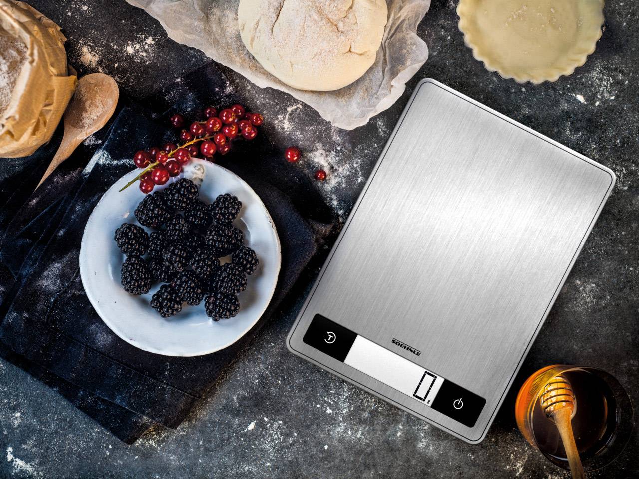 A stainless steel kitchen scale shows '0' grams. Beside it are berries, dough, flour, a wooden spoon, honey, and a baking tin on a flour-dusted surface.