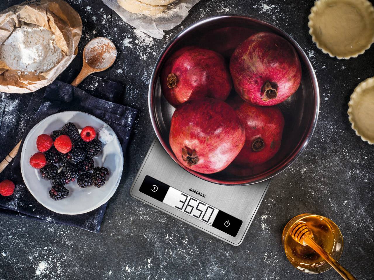 A kitchen scales shows 3,650 grams. Four pomegranates are placed in a bowl on the scales. Beside them are berries and honey.