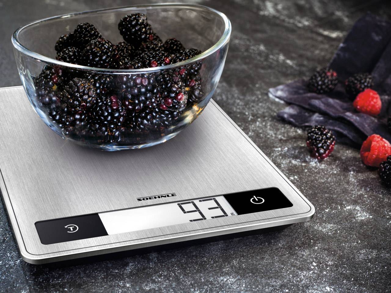 A glass bowl with blackberries sits on a digital kitchen scale, which displays 93 grams. Several more berries are scattered nearby.
