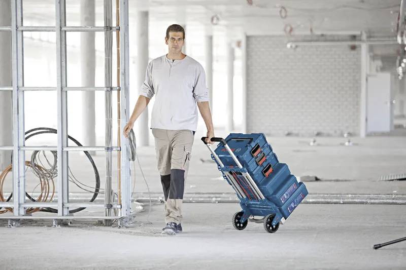 A worker in casual attire pulls a blue hand truck with stacked toolboxes across a large, unfinished construction site.