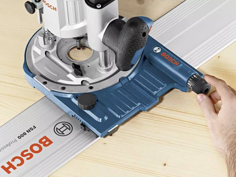 A hand guides a Bosch router along a guide rail on a wooden surface.