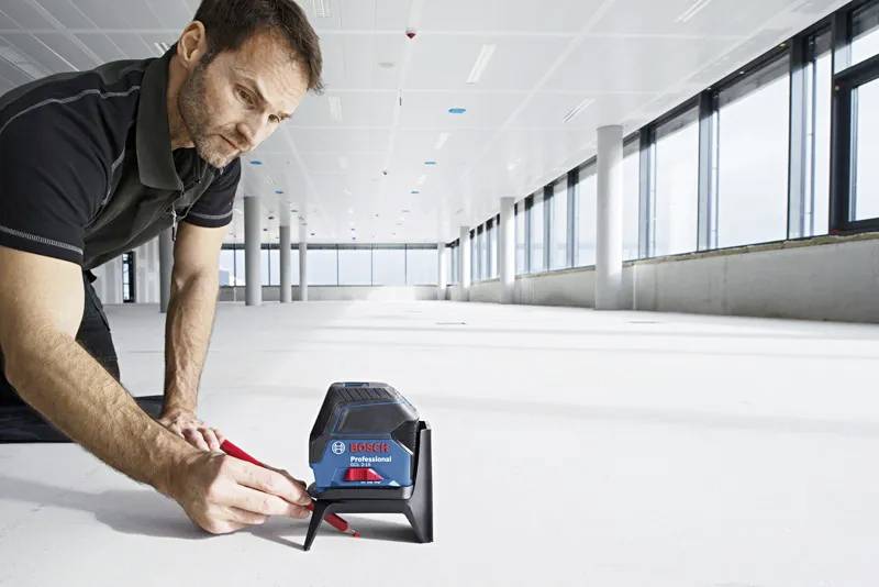 A person using a laser level tool on a large, empty floor to ensure alignment and precision in construction. Bright natural light fills the space.