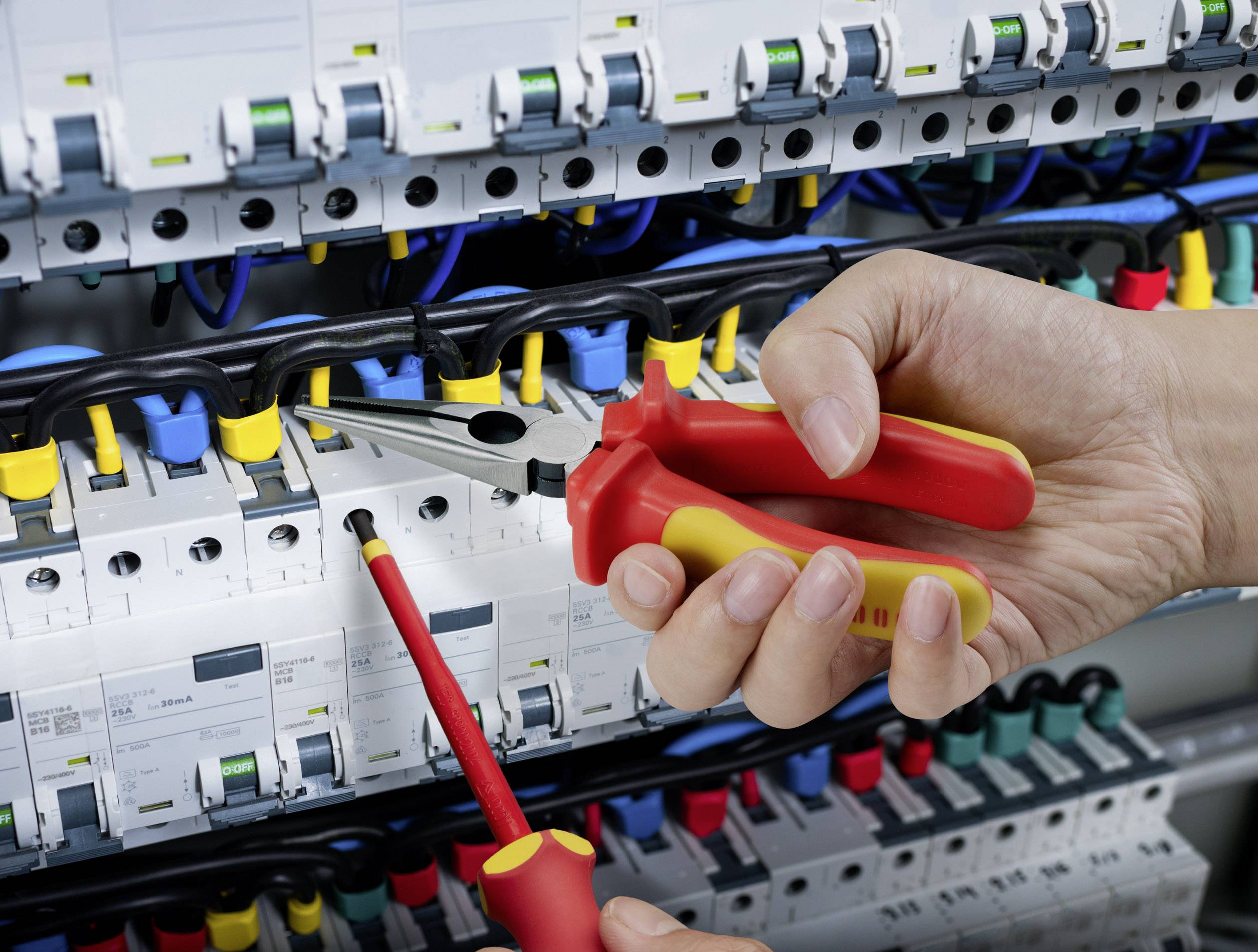 A hand holds pliers and a screwdriver in front of an open electrical cabinet full of cables and fuses that are being serviced.