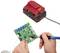 'A person is soldering a green circuit board that is connected to a red charger. Technology and repair in focus.'
