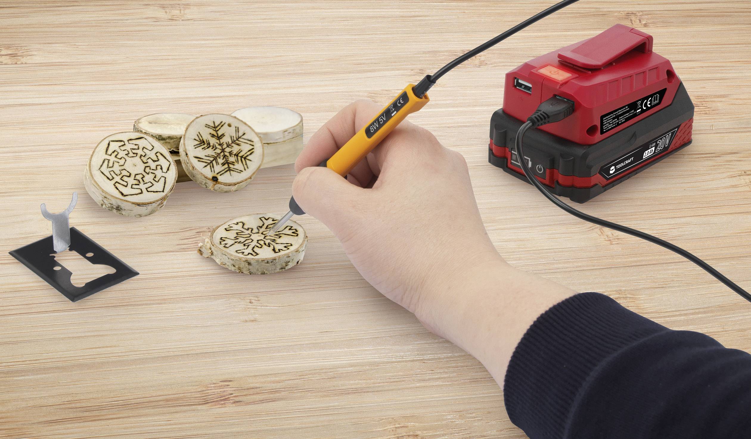 A hand is using a pyrography tool to burn patterns into round wooden discs. Beside it is a red and black equipment set.